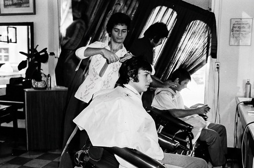 A young Eli cutting a client's hair on a busy day in a checkerboard-floor barbershop