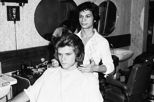 Eli and a fellow barber cutting a young man's hair beneath round wall mirrors
