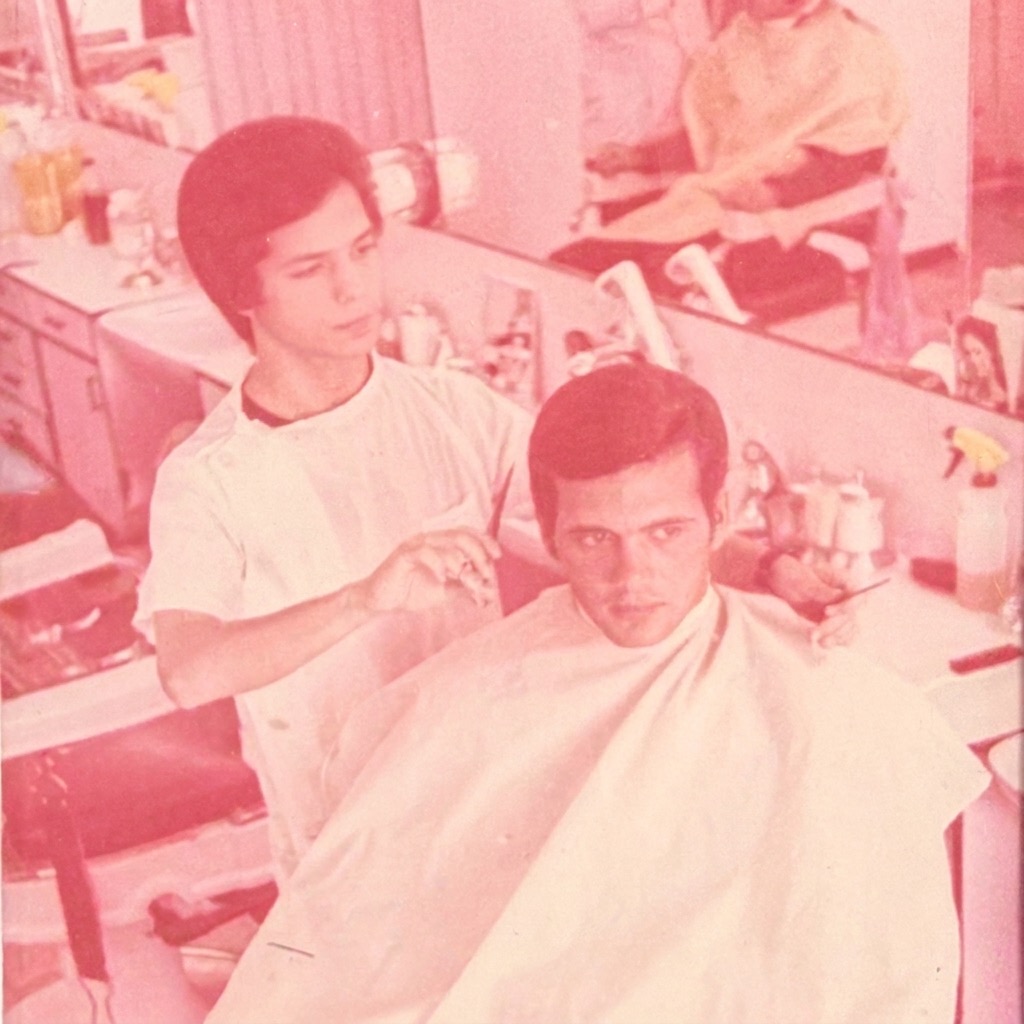 A young Eli cutting a client's hair in a vintage barbershop, reflected in the mirror behind him